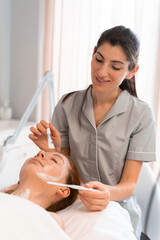 Wall Mural - Happy massage therapist applying face mask to woman with brush at cosmetology salon. Relaxed female customer is getting pampered in spa by professional