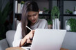© Wanda - Asian woman sits on sofa using digitalized pen writing on tablet with laptop on table in living room