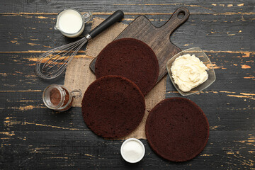 Canvas Print - Delicious chocolate sponge cake cut into layers with ingredients and whisk on dark wooden background