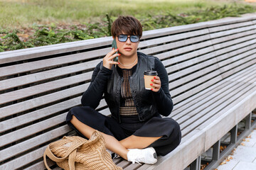 Brunette with short hair, sitting on a bench in the lotus position in a city park, holding a phone, talking, gesticulating, drinking coffee from a cardboard cup, on a summer day.