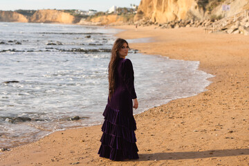 Wall Mural - beautiful young woman dancing flamenco walks along the shore of the beach at sunset, wearing a purple velvet dress with ruffles.