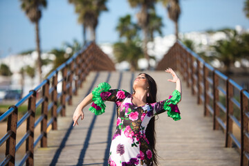 Wall Mural - Latin woman, brunette, with flowing hair, dancing flamenco on a wooden catwalk by the seashore. The woman is wearing a white dress with a floral print and ruffles. Intangible Cultural Heritage.