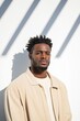 © Iryna - A man with dreadlocks stands in front of a white wall. He is wearing a tan jacket and a white shirt
