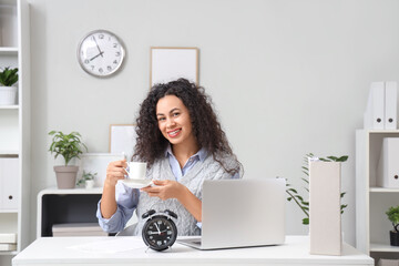 Wall Mural - Young African-American businesswoman with coffee cup at table in office. Time management concept