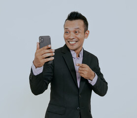 Wall Mural - Excited asian businessman smiling while pointing to mobile phone that he hold, isolated over white background.