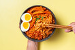 © Daniel - A bowl of spicy Korean ramen with red broth, topped with soft-boiled eggs, fish cakes, and garnished with green onions. A hand holds the bowl while another hand uses chopsticks to pick up the noodles