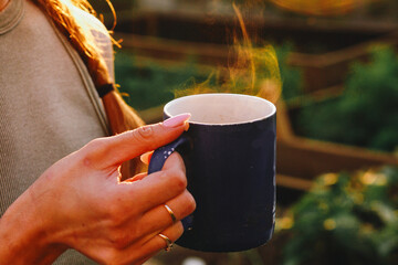 A girl with rings on her fingers holds a mug with a hot drink in her hands. Steam comes from a mug, tea party in the evening at sunset during the golden hour