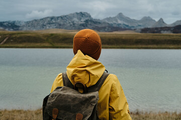 A young traveler woman wearing yellow raincoat and retro style backpack, hiking in mountains and enjoying views of nature. Rear view. Vrazje jezero - Montenegro country, Durmitor national park.