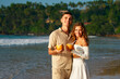 © artiemedvedev - Romantic couple enjoys sunset on tropical beach with drinks. Smiling man and woman toast coconuts, date by sea. Warm light on happy lovers. Casual summer wear, honeymoon vibes, love concept.