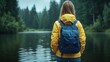 © imagineRbc - A person wearing a bright yellow jacket and carrying a backpack overlooks a serene lake. The backdrop features dense evergreen trees under a grey sky, creating a peaceful atmosphere