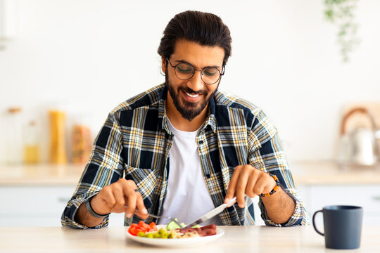 Positive arab guy hipster having healthy breakfast at home, eating fresh vegetables, sausages, drinking coffee, sitting at kitchen table, copy space. Handsome indian man starting day with nice meal