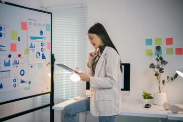 Wall Mural - Professional asian businesswoman analyzing financial data on clipboard, referencing whiteboard charts in contemporary workspace