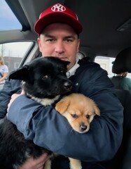 Man holds two puppies in his arms while inside of a car wearing red cap