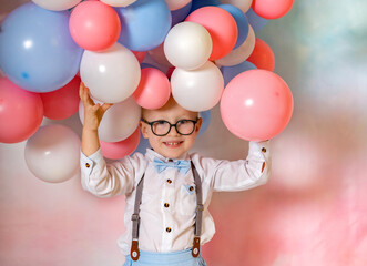 Cute little boy wearing birthday glasses and holding blue and pink balloons on a colored wall background