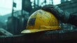 © Lucianastudio - A worn yellow hard hat rests on a dark surface a gloved hand gently placed on it in a industrial scene
