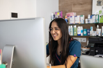 young veterinarian smiling and working on desktop computer in a veterinary clinic with shelves full 
