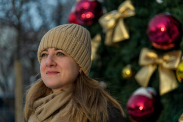 Wall Mural - A young woman wearing a knitted hat and scarf stands thoughtfully by a Christmas tree adorned with red ornaments and golden bows, basking in the holiday spirit.