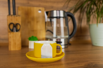 A yellow cup of tea stands on a yellow saucer and on the saucer lies a tea bag on a wooden table against the background of a teapot, a plant and kitchen utensils. The concept of tea drinking, break