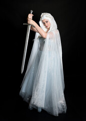 Wall Mural - full length portrait beautiful blonde fantasy maiden faery wearing medieval fairytale ballgown costume, flowing white hooded cape. standing pose holding a sword weapon, isolated dark studio background