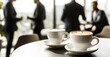 © David Giraud - Une réunion professionnelle avec des tasses de café sur un table, arrière-plan flou avec des gens en costume debout, un espace de bureau avec de grandes fenêtres donnant sur l'extérieur.