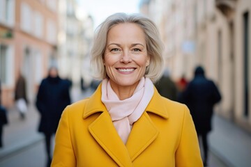 Wall Mural - Mature woman with cheerful smile stands in city street. Stylish in bright yellow coat, light pink scarf. Urban scene busy. Looks happy, confident. Outdoors in winter. Image embodies blend of urban