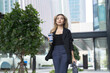© ultramansk - A confident young professional woman walking outdoors in a modern urban setting, holding a coffee cup, surrounded by greenery and glass buildings, exuding elegance and contemporary style.