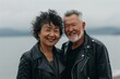 © Markus Schröder - Portrait of a joyful multicultural couple in their 50s sporting a classic leather jacket isolated on tranquil ocean backdrop