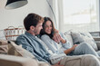 © Daniel - Young happy couple having fun talking laughing relaxing at home on couch, boyfriend embracing girlfriend telling funny joke sitting on sofa, humor in relationships, enjoying weekend together.