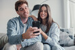 © Daniel - Cute couple greeting having fun video calling long distance friends and family catching up..Young man holding phone with girlfriend sitting on the sofa laughing and enjoying technology