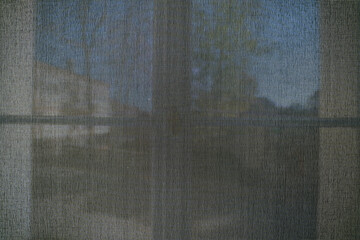 Close-up of the texture of a mosquito net on a window, through which a blurred background with houses and the sky can be seen.