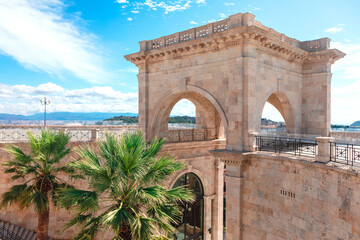 Bastione di Saint Remy in Cagliari, Sardinia Italy. Historic stone archway structure with two large arches, surrounded by palm trees and a scenic view of the sea and mountains in the background