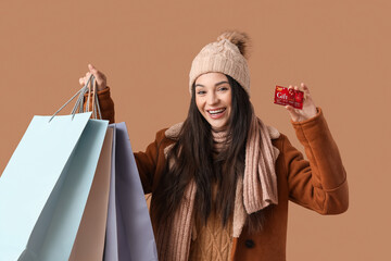 Poster - Beautiful young woman in warm clothes with gift card and shopping bags on brown background