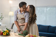 © crizzystudio - Young asian man wearing an apron is cutting fresh vegetables while his wife lovingly kisses him on the cheek in a bright, modern kitchen, enjoying their time together