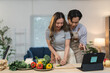 © crizzystudio - Happy asian couple cooking together in kitchen, woman slicing cucumber, man pointing at digital tablet following online recipe, fresh vegetables on table