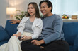 © crizzystudio - Happy asian couple laughing and watching a funny movie on television while sitting together on a comfortable sofa in their living room, enjoying their leisure time