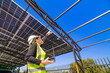 © ultramansk - A female engineer in a safety vest and helmet inspecting a solar carport under construction. The structure’s steel beams and partially installed solar panels are visible under a bright blue sky.