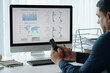 © DragonImages - Person monitoring business performance on computer screen with displayed charts and graphs, using smartphone. Organized workspace with office supplies in background