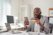 © Tj - Smiling African American businessman holding a credit card while using a smartphone for seamless online shopping at his modern workplace, enjoying the convenience of digital transactions