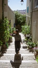 Wall Mural - Woman strolling cobblestone street in european city surrounded by lush plants and historical architecture under the bright sun.