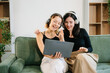 © Nuttapong punna - Two cheerful women sitting on a green couch, sharing ideas using a laptop and phone in a stylish home setup.
