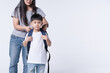 © bonnontawat - Asian mother assisting her confident young son with his backpack, preparing him for school, isolated in a studio.