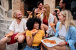 © CarlosBarquero - Group of cheerful young multiethnic friends are laughing and enjoying eating street food pizza together while sitting on steps in front of a historical building