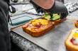 © pavel siamionov - A close-up of a chef's hands assembling a gourmet sandwich on toasted bread. Layers of bacon, cheese, and fresh cucumbers are being added. The kitchen setting is modern and inviting.