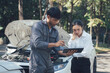 © Kritdanai - Car Trouble Consultation: A concerned female customer discusses car repair options with a professional male mechanic using a digital tablet, standing by a vehicle with its hood open.