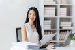© Kritdanai - Confident Businesswoman at Desk: A young, smiling businesswoman sits at her modern office desk, reviewing documents.  She exudes confidence and competence.