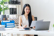 © Kritdanai - Focused and Efficient: A young businesswoman with a confident smile, analyzes financial documents at her desk, surrounded by an organized workspace. The image exudes efficiency and dedication.