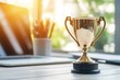 © Alexandr - Golden trophy on an office desk alongside a laptop and potted plant, representing achievement, success, and motivation in a professional setting