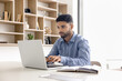 © fizkes - Young 20s Arabian man sitting at workplace desk, holding pen and typing on laptop, taking important notes while researching online, editing document, update report or spreadsheet, responding to emails