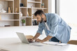© fizkes - Young 30s businessman, individual entrepreneur standing leaned at workplace desk working on laptop, search information, do remote telework use modern platform, internet connection. Deadline, workflow