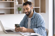 © fizkes - Smiling Latino man sit at workplace desk with laptop, distracted from online work, using modern smartphone, enjoy personal chat, check information, browse website, download new business application